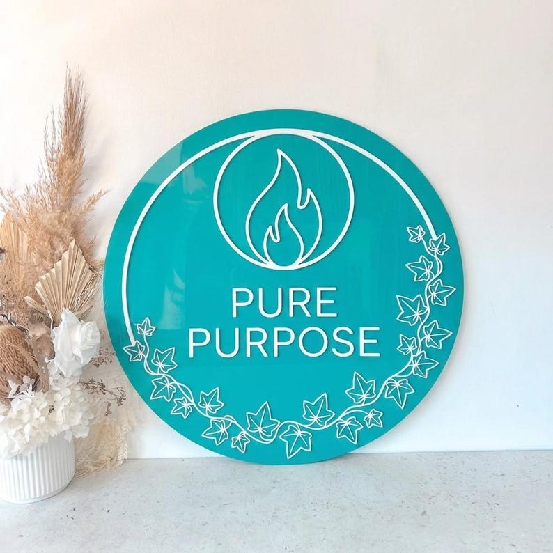 May include: A round teal sign with the words "PURE PURPOSE" in white. A white flame graphic is centered above the text, within a white circle. White ivy leaf designs border the bottom and right side of the sign.