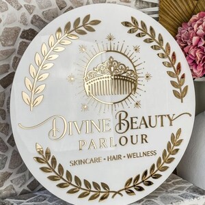 May include: A round white sign with gold lettering and accents. The sign reads "Divine Beauty Parlour" and includes the words "Skincare, Hair, Wellness." A gold comb and crown design is in the center, surrounded by a sunburst and laurel leaves.
