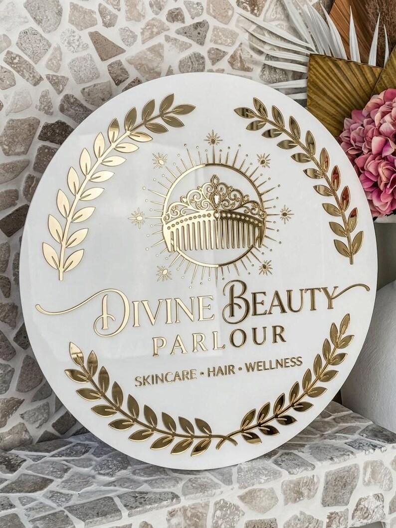 May include: A round white sign with gold lettering and accents. The sign reads "Divine Beauty Parlour" and includes the words "Skincare, Hair, Wellness." A gold comb and crown design is in the center, surrounded by a sunburst and laurel leaves.