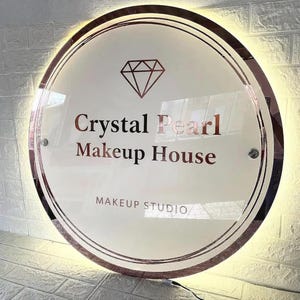 May include: A circular, illuminated sign with the words "Crystal Pearl Makeup House" in rose gold. The sign includes a diamond graphic and the text "Makeup Studio". It has a rose gold border and is mounted on a wall.