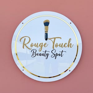 May include: A round white sign with gold trim and the words "Rouge Touch Beauty Spot" in gold and navy blue script. A makeup brush graphic is at the top. The sign is mounted on a pink wall.