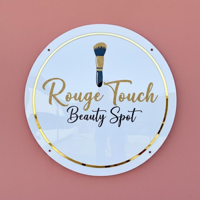 May include: A round white sign with gold trim and the words "Rouge Touch Beauty Spot" in gold and navy blue script. A makeup brush graphic is at the top. The sign is mounted on a pink wall.