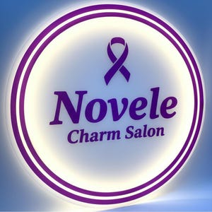 K&ouml;nnte beinhalten: Rundes, beleuchtetes Schild mit einer lila Schleife und dem Schriftzug "Novele Charm Salon" in Lila. Das Schild hat eine wei&szlig;e Mitte, einen lila Rand und einen blauen Farbverlauf im Hintergrund.
