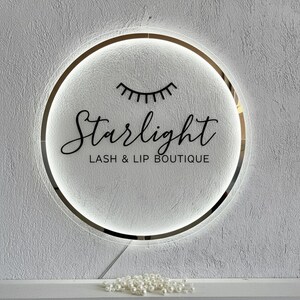 Pu&ograve; includere: Un'insegna rotonda e illuminata con la scritta "Starlight LASH & LIP BOUTIQUE" in elegante corsivo. L'insegna presenta un bordo dorato e un design stilizzato di ciglia. Lo sfondo &egrave; un muro bianco strutturato.