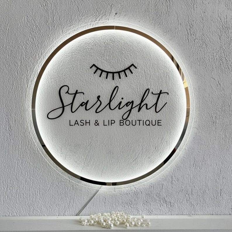 Pu&ograve; includere: Un'insegna rotonda e illuminata con la scritta "Starlight LASH & LIP BOUTIQUE" in elegante corsivo. L'insegna presenta un bordo dorato e un design stilizzato di ciglia. Lo sfondo &egrave; un muro bianco strutturato.