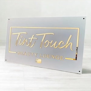 May include: A rectangular sign with the words "Tint Touch BEAUTY LOUNGE" in gold lettering. The sign has a light gray background with a gold border and a small gold graphic of a brush.