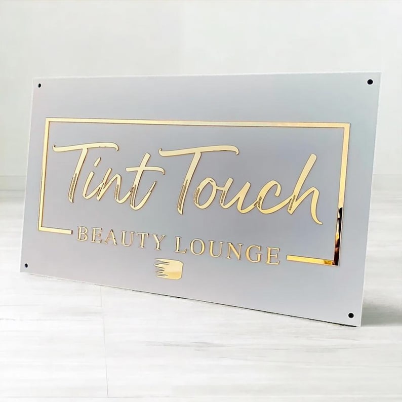 May include: A rectangular sign with the words "Tint Touch BEAUTY LOUNGE" in gold lettering. The sign has a light gray background with a gold border and a small gold graphic of a brush.