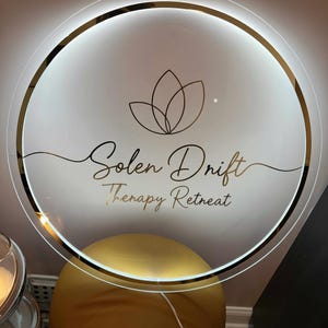 Könnte beinhalten: Ein rundes, beleuchtetes Schild mit goldenem Rand und den Worten "Solen Drift Therapy Retreat" in eleganter Schrift. Das Schild zeigt ein goldenes Lotusblumen-Design und wird von einem sanften, weißen Schein beleuchtet.