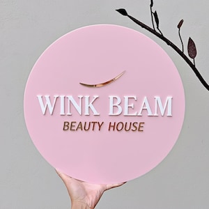 May include: A round, pink sign with the words "WINK BEAM BEAUTY HOUSE" in white and gold lettering. A gold eyelash design is above the text. The sign is held by a hand.