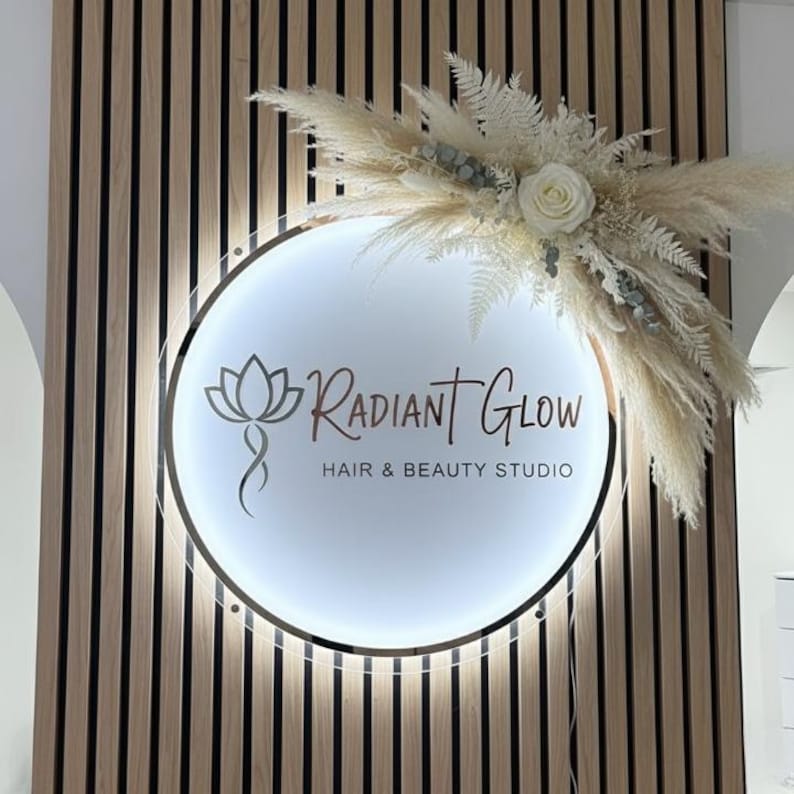 May include: A circular, illuminated sign for "Radiant Glow Hair & Beauty Studio", featuring a floral design and text. The sign is adorned with dried flowers and pampas grass, mounted on a wooden panelled wall.