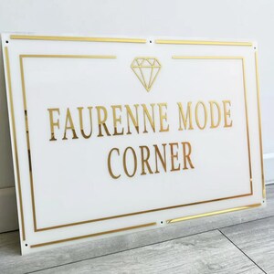 May include: White rectangular sign with gold trim and lettering. The sign reads "FAURENNE MODE CORNER" in gold, with a gold diamond graphic above the text. The sign is mounted on a wall.