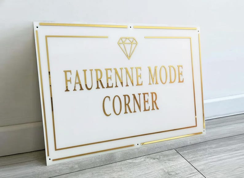 May include: White rectangular sign with gold trim and lettering. The sign reads "FAURENNE MODE CORNER" in gold, with a gold diamond graphic above the text. The sign is mounted on a wall.