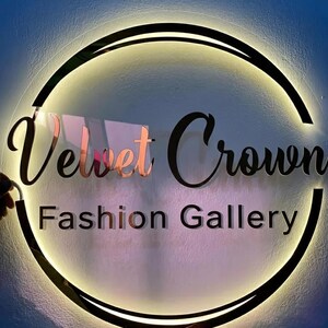 K&ouml;nnte beinhalten: Rundes Schild mit den Worten "Velvet Crown Fashion Gallery" in einer stilvollen Schriftart. Das Schild hat einen schwarzen Rand mit einem warmen, leuchtenden Licht am Rand. Die W&ouml;rter "Velvet" und "Crown" sind ros&eacute;goldfarben.