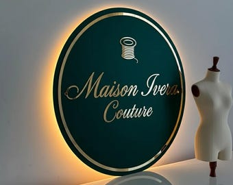 May include: A teal and gold illuminated sign with the words "Maison Ivera Couture" and a spool icon. A dress form stands to the right. The sign is oval-shaped with a gold border and a warm glow.