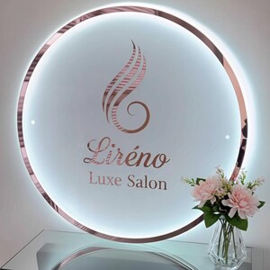 K&ouml;nnte beinhalten: Ein runder, beleuchteter Schriftzug mit ros&eacute;goldfarbenem Rahmen und dem Text "Lir&eacute;no Luxe Salon". Das Schild zeigt ein stilisiertes Federdesign in Ros&eacute;gold. Unter dem Schild steht eine kleine Vase mit rosa Blumen.