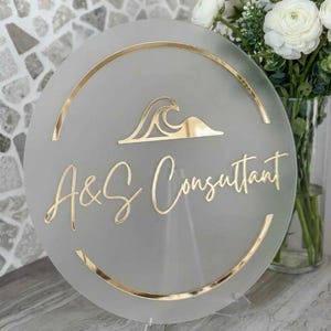 May include: A round, frosted glass sign with gold accents and lettering. The sign features the text "A&S Consultant" in a stylish script, along with a gold wave design. The sign is displayed on a clear stand.