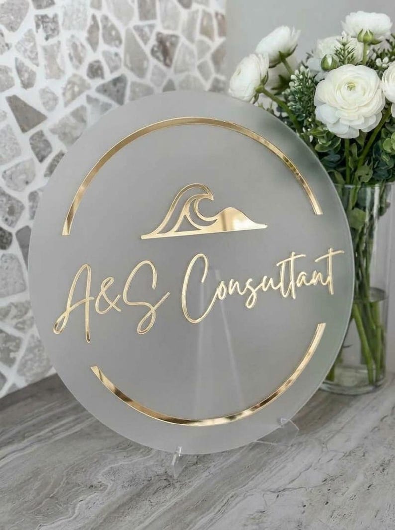 May include: A round, frosted glass sign with gold accents and lettering. The sign features the text "A&S Consultant" in a stylish script, along with a gold wave design. The sign is displayed on a clear stand.