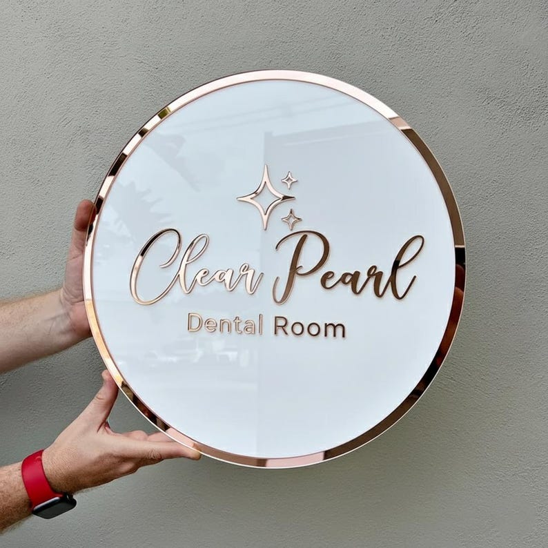 May include: A round, white sign with a rose gold border and lettering. The sign reads "Clear Pearl Dental Room" in elegant script. Above the text are two starburst designs. The sign is held by a person.