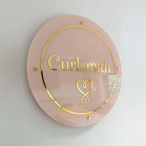 Pu&ograve; includere: Un cartello rotondo rosa con la scritta dorata "Curlsmith". Il cartello presenta un disegno a forma di cuore dorato e un bordo dorato. Tre piccoli punti dorati sono posizionati attorno al perimetro del cartello.