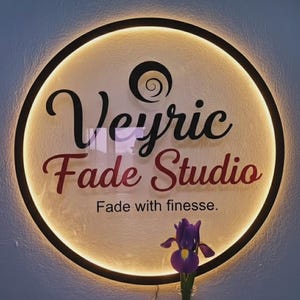 Puede incluir: Un letrero redondo e iluminado con las palabras "Veyric Fade Studio" en escritura negra y burdeos. El letrero tiene un borde negro y un brillo suave y c&aacute;lido. Debajo del texto est&aacute; la frase "Fade with finesse".
