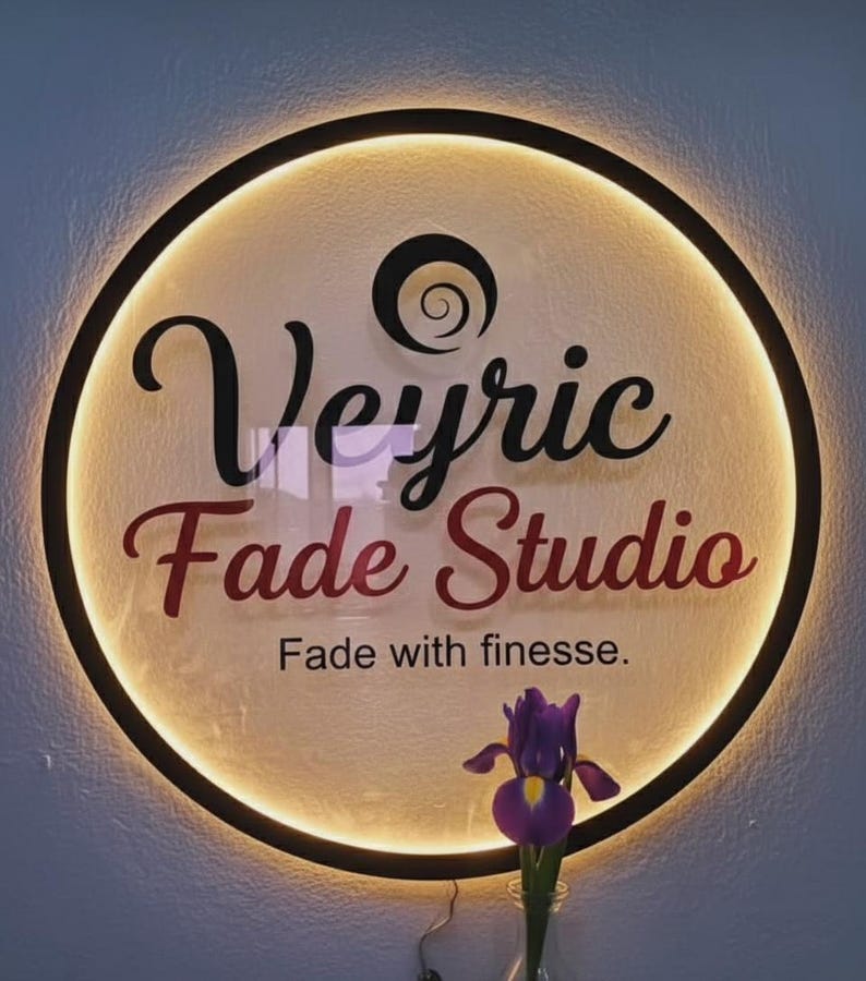 Puede incluir: Un letrero redondo e iluminado con las palabras "Veyric Fade Studio" en escritura negra y burdeos. El letrero tiene un borde negro y un brillo suave y c&aacute;lido. Debajo del texto est&aacute; la frase "Fade with finesse".