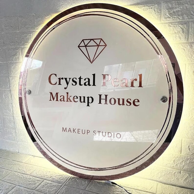 May include: A round, illuminated sign with the text "Crystal Pearl Makeup House" in a rose gold color. The sign also features a diamond graphic and the words "Makeup Studio". The sign is surrounded by a rose gold border.