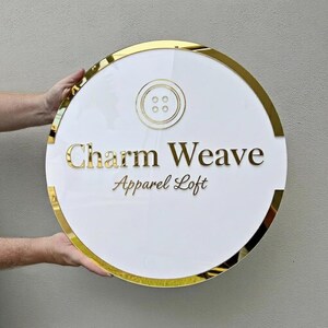 May include: A round, white sign with gold trim and lettering. The sign features the text "Charm Weave Apparel Loft" in gold, with a gold button graphic at the top. The sign is held by two hands.