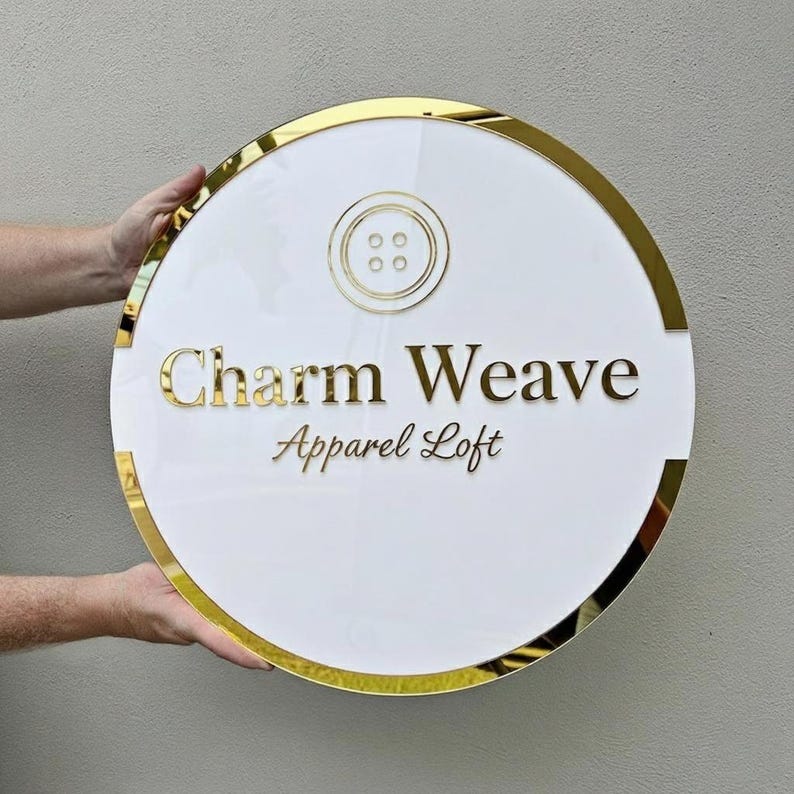 May include: A round, white sign with gold trim and lettering. The sign features the text "Charm Weave Apparel Loft" in gold, with a gold button graphic at the top. The sign is held by two hands.