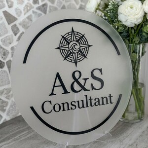 May include: A round, frosted glass sign with a black compass rose design and the text "A&S Consultant." The sign is displayed on a clear stand, with a bouquet of white flowers in the background.