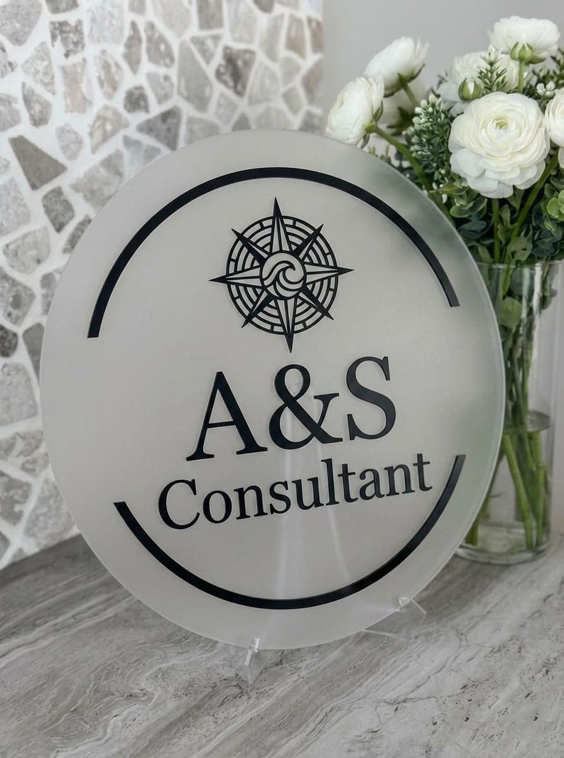 May include: A round, frosted glass sign with a black compass rose design and the text "A&S Consultant." The sign is displayed on a clear stand, with a bouquet of white flowers in the background.