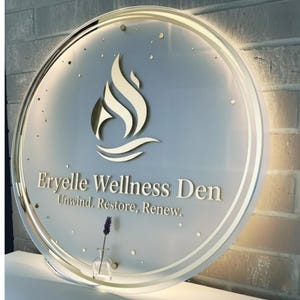 Puede incluir: Letrero circular para Eryelle Wellness Den con borde y logotipo dorados. El logotipo es una llama estilizada sobre l&iacute;neas onduladas. El letrero incluye el texto "Eryelle Wellness Den" y "Unwind. Restore. Renew."