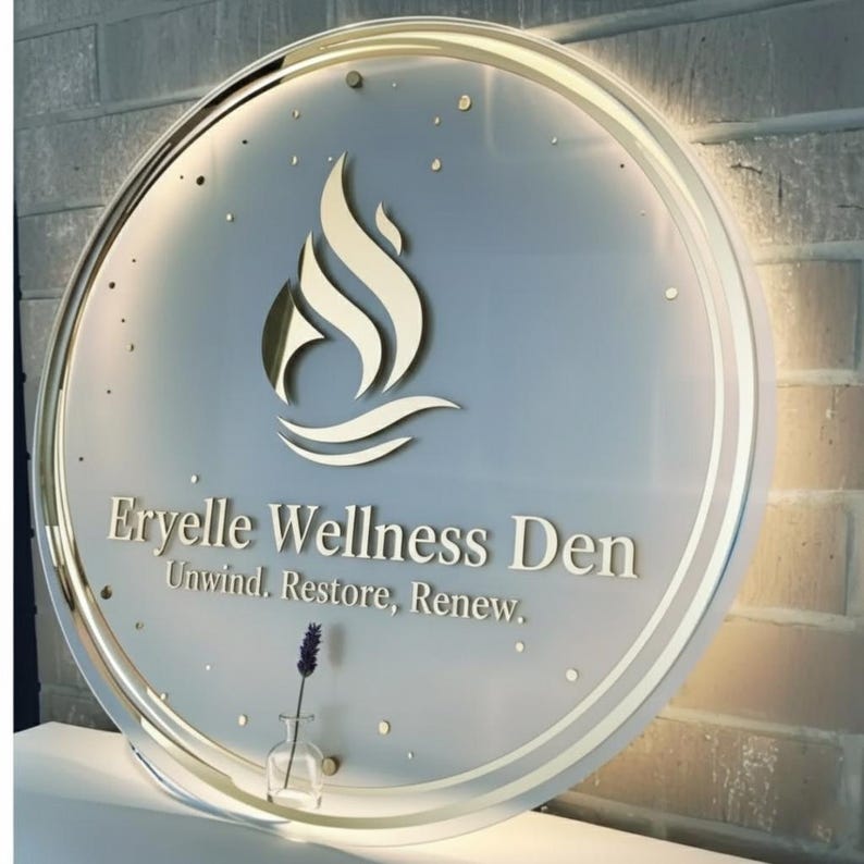 Puede incluir: Letrero circular para Eryelle Wellness Den con borde y logotipo dorados. El logotipo es una llama estilizada sobre l&iacute;neas onduladas. El letrero incluye el texto "Eryelle Wellness Den" y "Unwind. Restore. Renew."