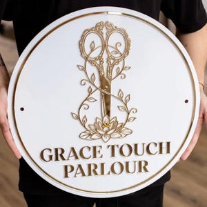 May include: A round white sign with gold trim and a gold scissors design. The sign reads "GRACE TOUCH PARLOUR" in gold lettering. The scissors are surrounded by floral accents.