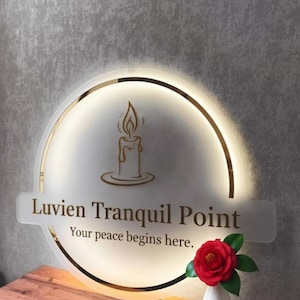 May include: A round, illuminated sign with a gold trim and the text "Luvien Tranquil Point" and "Your peace begins here." A candle graphic is in the center. A red rose in a white vase sits on a wooden surface.