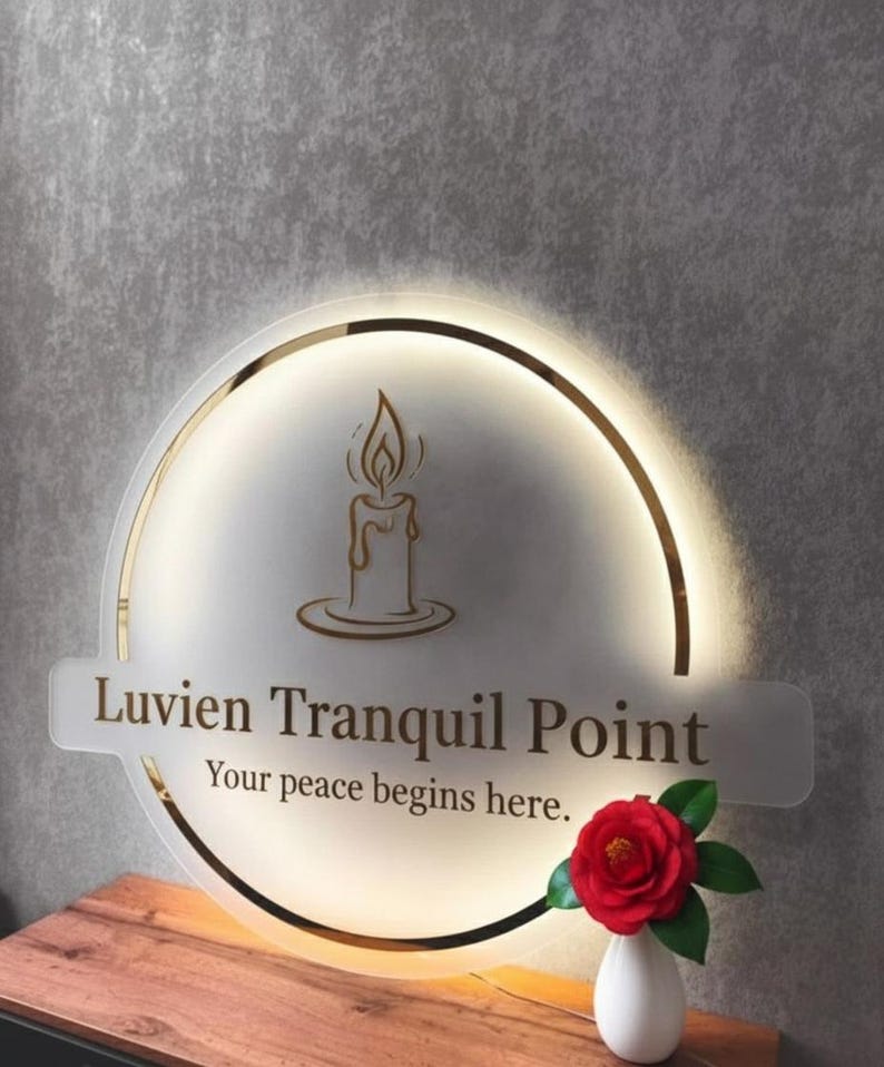 Puede incluir: Un letrero redondo e iluminado con borde dorado y el texto "Luvien Tranquil Point" y "Your peace begins here." Un gr&aacute;fico de vela est&aacute; en el centro. Una rosa roja en un jarr&oacute;n blanco est&aacute; sobre una superficie de madera.