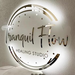 May include: A circular, illuminated sign displaying "Tranquil Flow Healing Studio" in a mirrored, cursive font. The sign has a frosted glass effect with a bright, white light behind the lettering and a decorative, mirrored border.