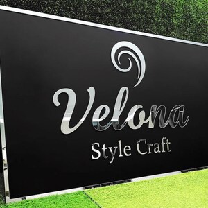 May include: A rectangular black sign with a silver mirrored border and lettering. The sign reads "Velona Style Craft" in a mirrored, elegant font. A decorative swirl is above the word "Velona". The sign is displayed on a green grassy surface.
