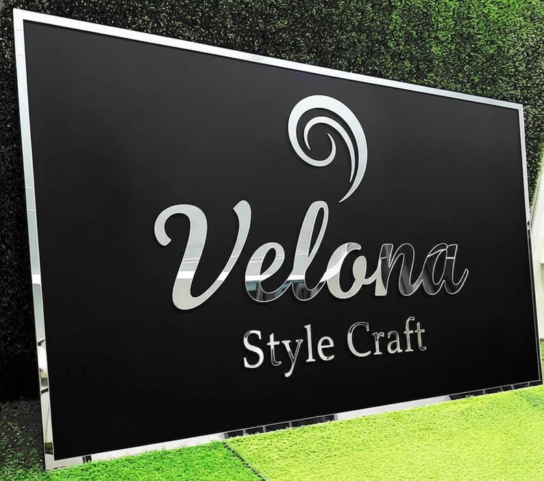 May include: A rectangular black sign with a silver mirrored border and lettering. The sign reads "Velona Style Craft" in a mirrored, elegant font. A decorative swirl is above the word "Velona". The sign is displayed on a green grassy surface.