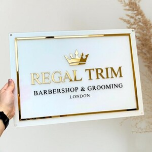 May include: A rectangular sign with a white background and gold trim. The sign reads "REGAL TRIM BARBERSHOP & GROOMING LONDON" in gold and black lettering, with a gold crown above the text. The sign is held by a person.