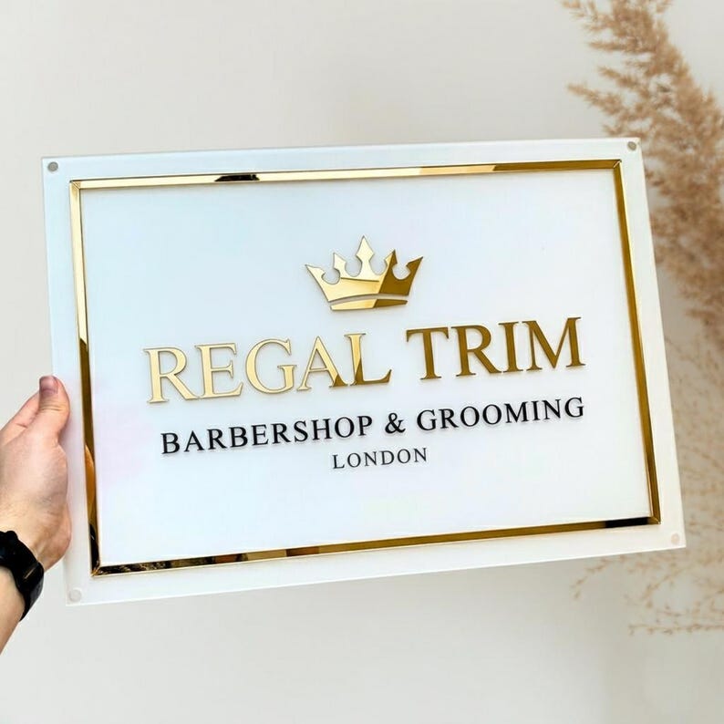 May include: A rectangular sign with a white background and gold trim. The sign reads "REGAL TRIM BARBERSHOP & GROOMING LONDON" in gold and black lettering, with a gold crown above the text. The sign is held by a person.