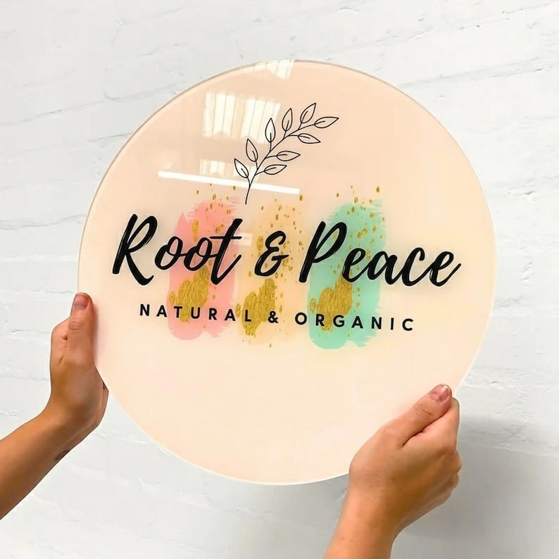 Pu&ograve; includere: Un cartello rotondo con le parole "Root & Peace" in corsivo nero, con "NATURAL & ORGANIC" sotto. Il cartello presenta pennellate rosa, oro e verde acqua e un rametto di foglie. Il cartello &egrave; tenuto da due mani.