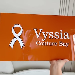 May include: An orange rectangular sign with the text "Vyssia Couture Bay" in white. A white ribbon graphic is on the left. The sign is being held up by a hand.
