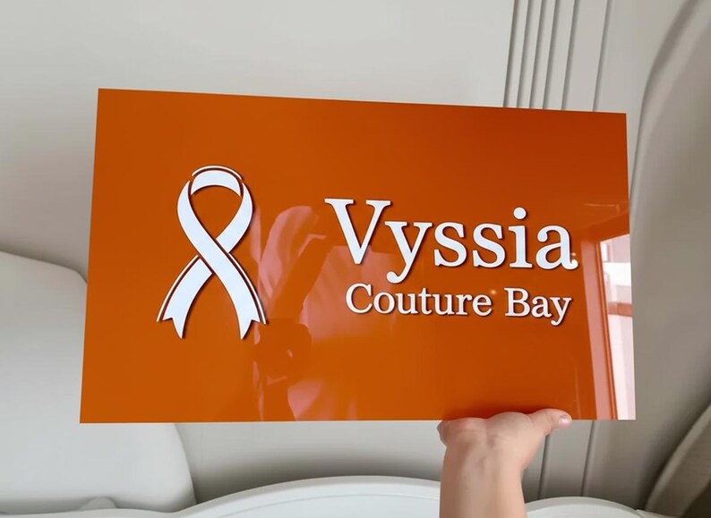 May include: An orange rectangular sign with the text "Vyssia Couture Bay" in white. A white ribbon graphic is on the left. The sign is being held up by a hand.