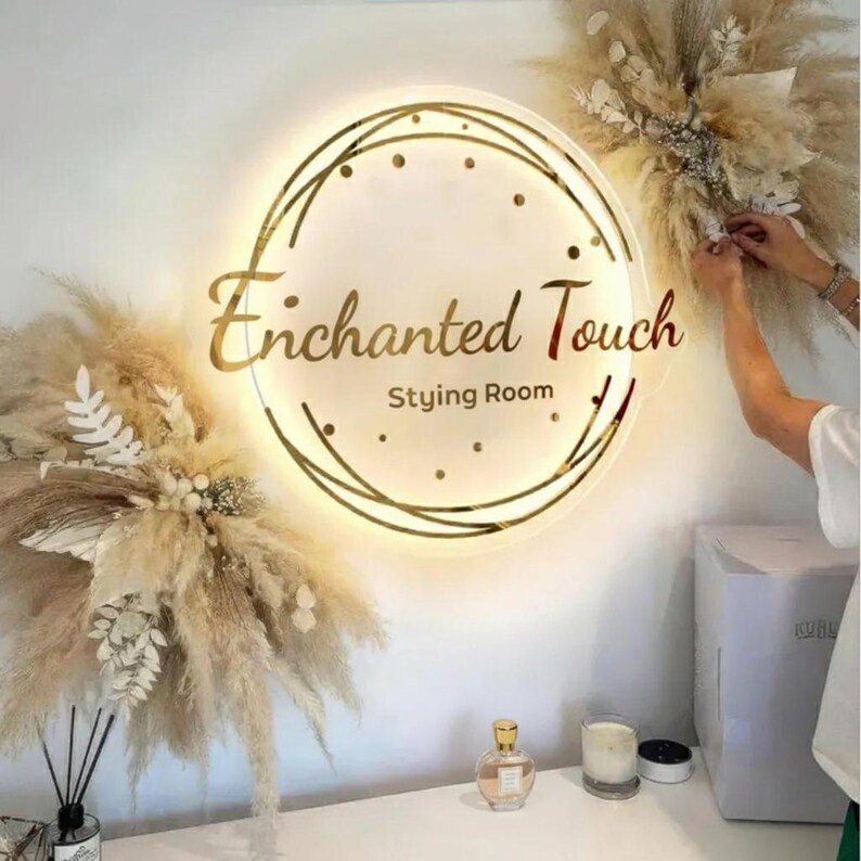 May include: A round illuminated sign with the words "Enchanted Touch Styling Room" in gold lettering. The sign is surrounded by a gold geometric design and small lights. Dried pampas grass and floral arrangements decorate the wall.