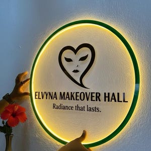 Puede incluir: Un letrero redondo e iluminado con un borde verde y un dise&ntilde;o en forma de coraz&oacute;n. El letrero dice "ELVYNA MAKEOVER HALL" y "Radiance that lasts." El letrero est&aacute; sostenido por dos manos.