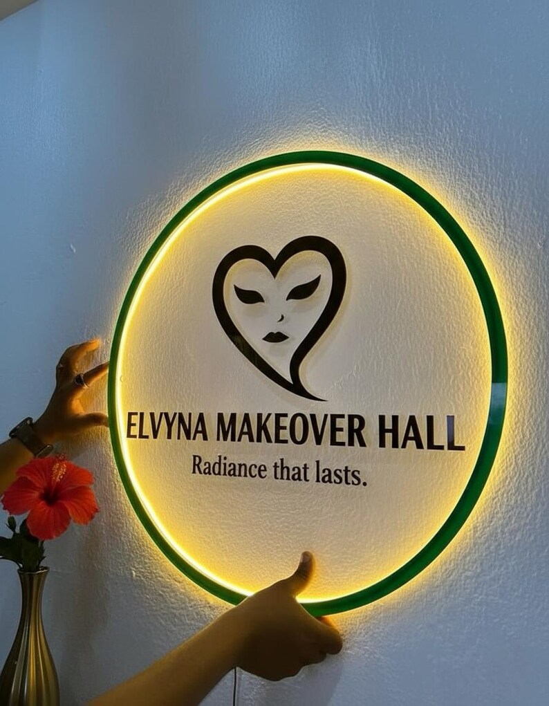 Puede incluir: Un letrero redondo e iluminado con un borde verde y un dise&ntilde;o en forma de coraz&oacute;n. El letrero dice "ELVYNA MAKEOVER HALL" y "Radiance that lasts." El letrero est&aacute; sostenido por dos manos.