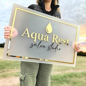 May include: A rectangular sign with a frosted glass center and gold trim. The sign reads "Aqua Rest salon Studio" in gold and black lettering, with a gold droplet symbol above the word "Rest".