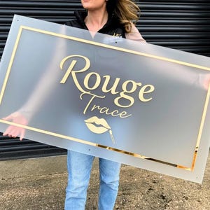 May include: A rectangular sign with the words "Rouge Trace" in gold script, a gold lip graphic, and a gold border. The sign is held up by a person wearing blue jeans.