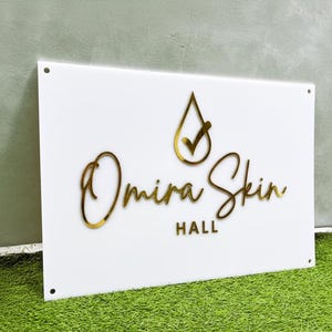 May include: A white rectangular sign with gold lettering and a gold logo. The sign reads "Omira Skin Hall". The logo is a gold teardrop with a checkmark. The sign has four holes in the corners.