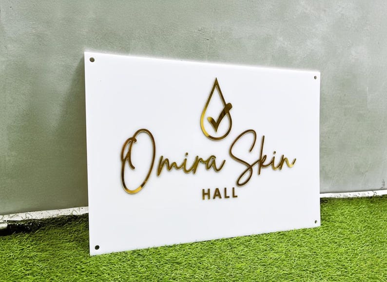 May include: A white rectangular sign with gold lettering and a gold logo. The sign reads "Omira Skin Hall". The logo is a gold teardrop with a checkmark. The sign has four holes in the corners.